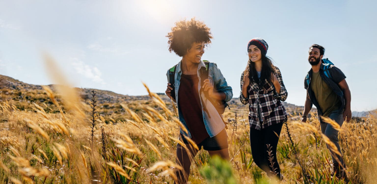 Three young adults with daypacks walking through a field of tall golden grass on a hillside, with the low sun behind them.