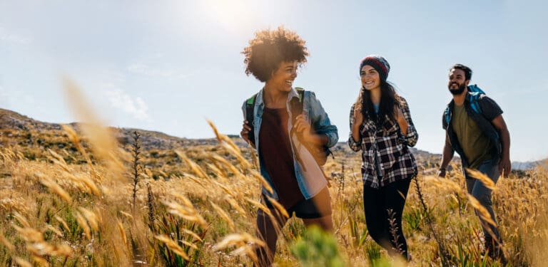 Three young adults with daypacks walking through a field of tall golden grass on a hillside, with the low sun behind them.