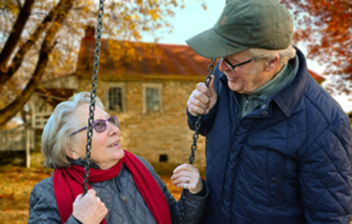 An older couple in winter coats smiling at each other beside a rope swing, with autumn leaves and a stone house behind them.