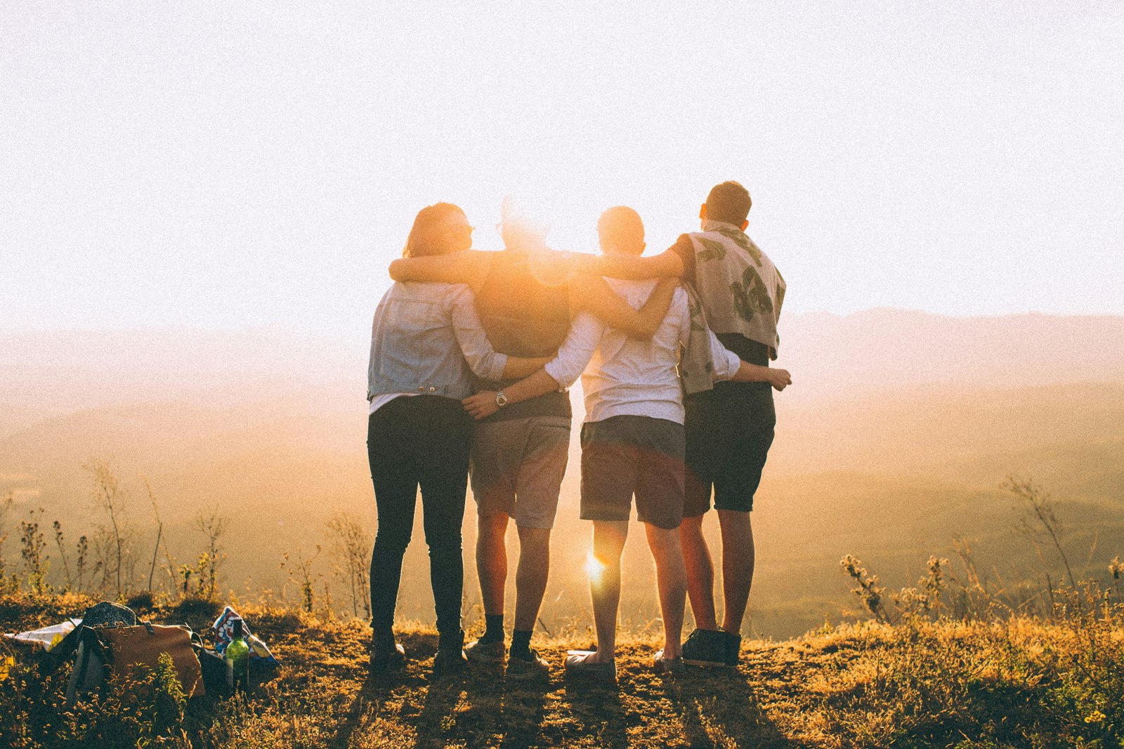 Four young adults standing arm in arm on a grassy hilltop, seen from behind as they look out at the sunset.